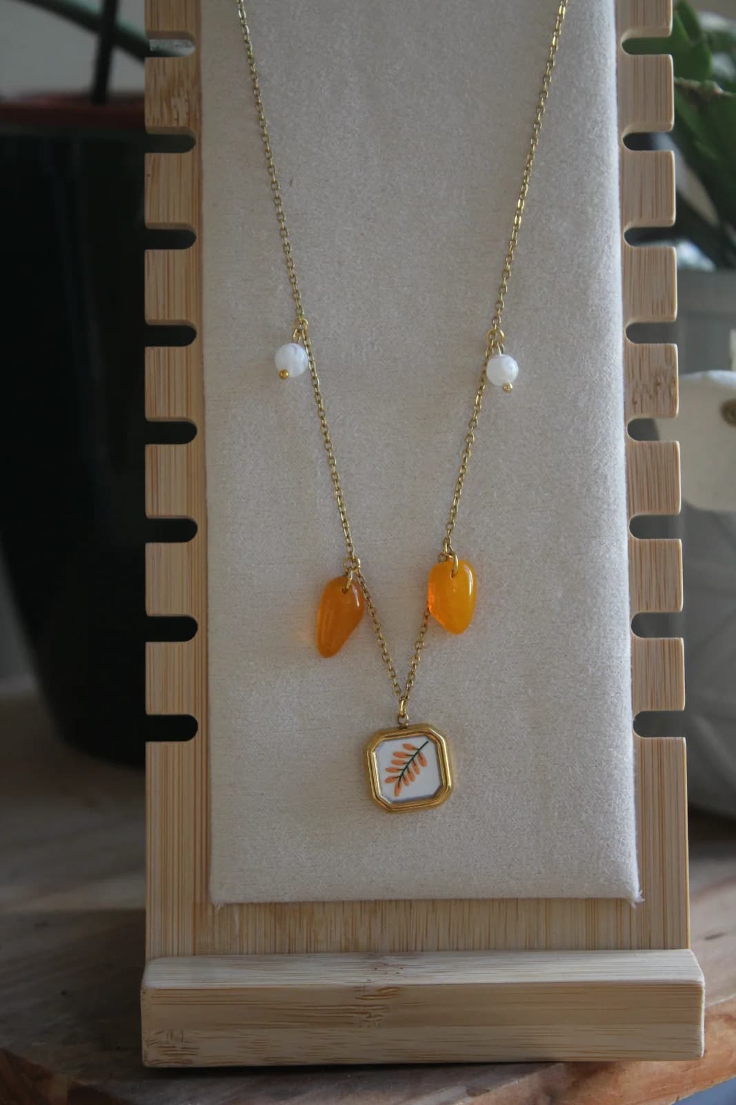 Collier doré à pendentif végétal, pampilles orange et perles blanches sur un présentoir en bois