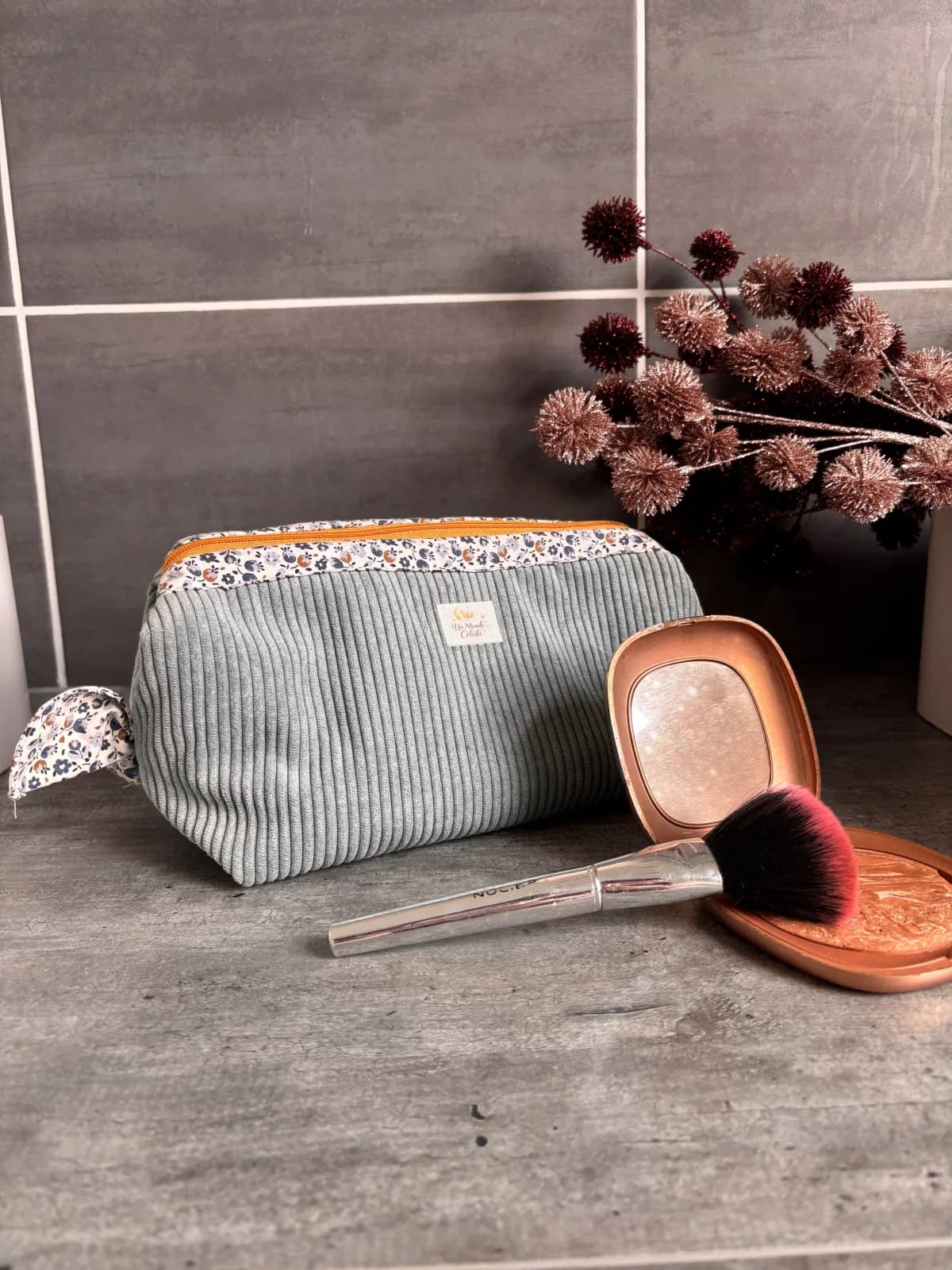 Trousse fermée en velours côtelé vert d'eau et tissu fleuri, posée près d'un pinceau de maquillage